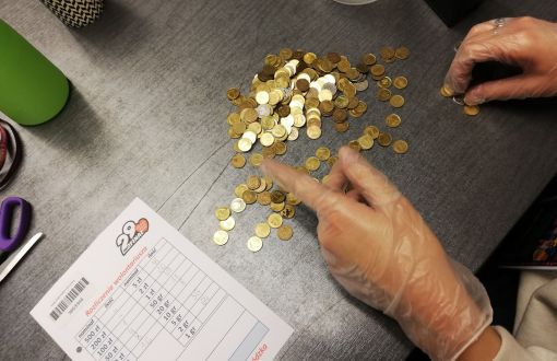 Na czarnym blacie złote monety i kartka do notowania. Widoczne ręce w przeźroczystych rękawiczkach.