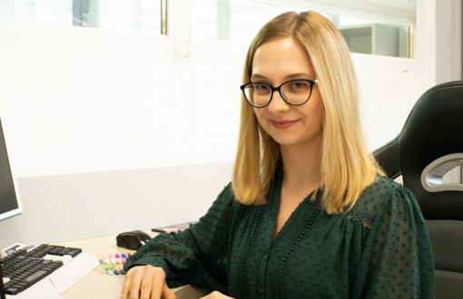 Zdjęcie portretowe: Monika Michałowska w ciemnej koszuli siedzi przy biurku i kompuerze.