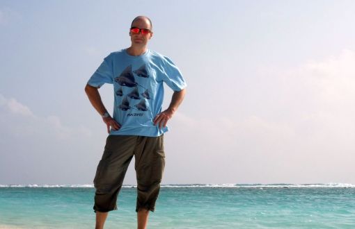 prof. K. Olejnik w przeciwsłonecznych okularach, niebieskim t-shircie i brązowych spodniach stoi na skraju morza.