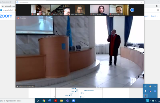 Zrzut z ekranu komputerowego: na górze siedem okien z uczestnikami spotkania. Poniżej, na 3/4 części strony podgląd z auli. Po lewej stronie katedra, po lewej stoły. Między nimi mężczyzna w todze.