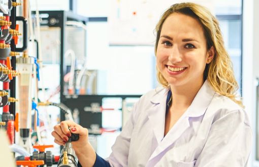 Uśmiechnięta studentka w białym fartuchu przy laboratoryjnej aparaturze.