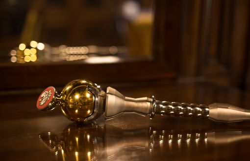 Srebrne berło PŁ ze złotymi elementami leży na blacie, w którym widoczne jest odbicie.