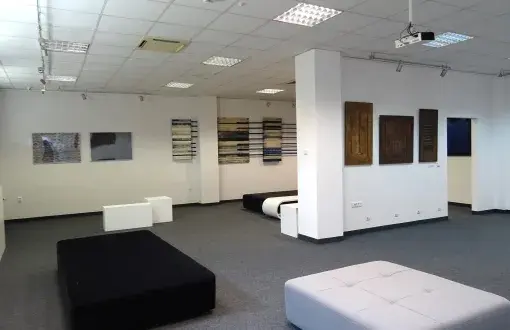 Pomieszczenie Galeria Biblio-Art: białe ściany, na nich prace graficzne. Na szarej wykładzinie stoją białe i czarne pufy.