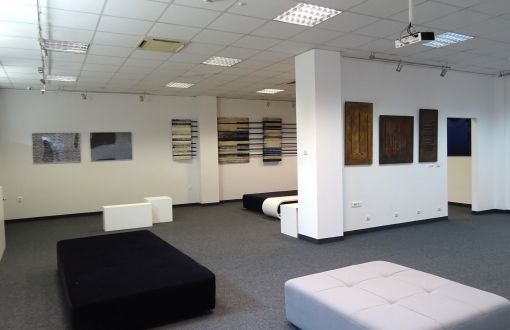 Pomieszczenie Galeria Biblio-Art: białe ściany, na nich prace graficzne. Na szarej wykładzinie stoją białe i czarne pufy.