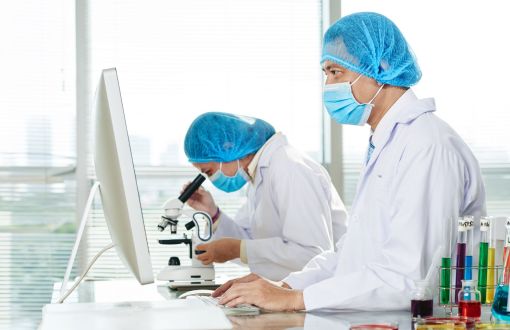 Chemical laboratory. Two researchers in white lab coats and blue lab caps and masks stand by a microscope and screen.