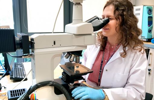 A young woman in a white lab coat looks through a microscope.