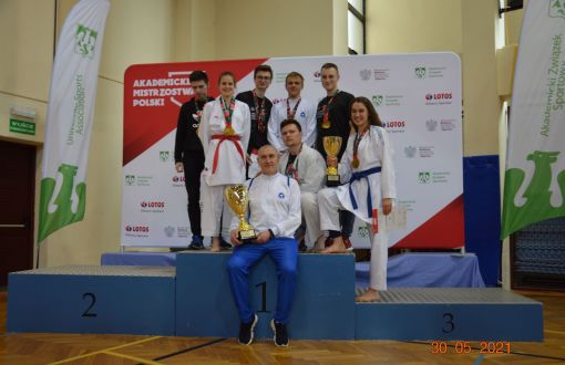 Duże niebieskie podium i laureaci Akademickich Mistrzostw Polski  w karate olimpijskim: centalnie siedzi trener z pucharem, w drugim rzędzie dwie zawodniczki w białych strojach i pięciu zawodników z medalami na szyjach.  W tle biało-czerwona ścianka reklamowa z napisem Akademickie Mistrzostwa Polski i logotypami sponsorów.