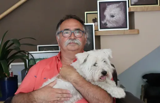Prof. Tomasz Kapitaniak w łososiowej polówce siedzi w fotelu i trzyma na rękach białego psa. W tle ściana ze zdjęciami tego samego psa.