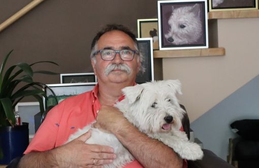 Prof. Tomasz Kapitaniak w łososiowej polówce siedzi w fotelu i trzyma na rękach białego psa. W tle ściana ze zdjęciami tego samego psa.