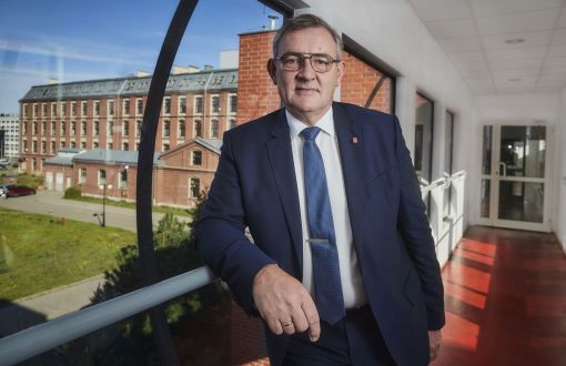 Prof. Krzysztof Jóźwik, rektor PŁ, stoi przeszklonym pomieszczeniu z widokiem na kampus uczelni i fragment ceglanego Budynku Trzech Wydziałów z regularnie rozłożonymi oknami.