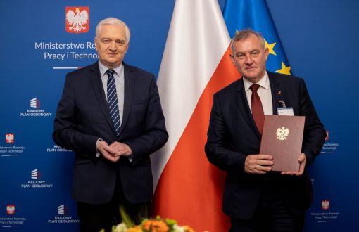 Na tle niebieskiej ścianki z napiem Ministerstwo Rozwoju, Pracy i Technologii oraz flag Unii Europejskiej i Polski stoją w garniturach: Jarosław Gowin (po lewej) i prof. Krzysztof Jóźwik (po prawej), który trzyma bordową teczkę ze złotym godłem Polski.