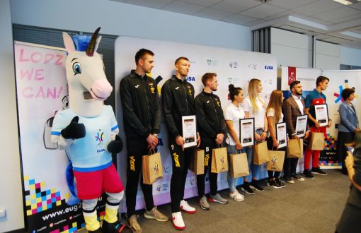 Od lewej strony, w jednym rzędzie stoją: maskotka EUG2022 - kolorowy jednorożec i ośmiu ambasadorów wydarzenia w strojach sportowych.