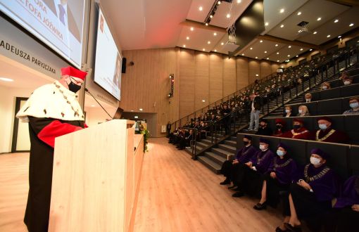 Zdjęcie auli z boku: po lewej stronie Rektor PŁ w todze stoi przy mównicy, po prawej stronie rzędy krzeseł.