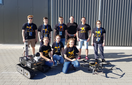 Członkowie drużyny Raptors z łazikiem i dronem na tle ciemnego budynku.