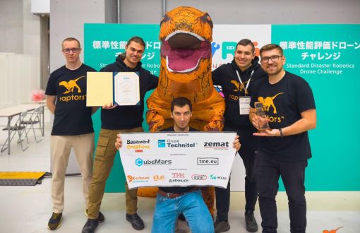 5 studentów z uśmiechem pozuje do zdjęcia stojąc wokół dużej figury dinazaura pomarańczowego