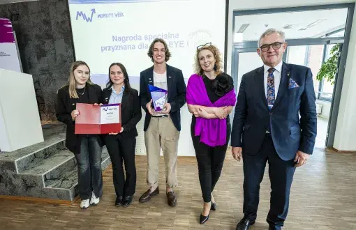 pięcioro ludzi stoi na tle wyświatlanego slajdu oraz szklanych dzwi, 2 trzymają  dyplom,  1 statuetkę, a 2 kolejne pozują, uśmiechając