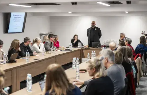 grupa osób siedzi przy ustawionych na przeciw siebie stołach, 1 mężczyzna stoi, jest ubrany na czarno