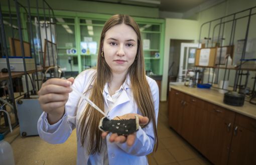 studentka w białym fartuchu trzyma pipetę oraz kawałek ciemnego materiału, na którym widać skroploną wodę, w tle zyblurowane laboratorium
