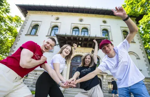 Czworo radosnych młodych osób z rękoma uniesionymi do góry. W tle zabytkowa willa i drzewa 