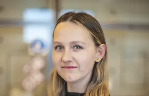 młoda kobieta w ciemnej bluzce, lewy profil, w tle rozmazane elementy wyposażenia laboratorium