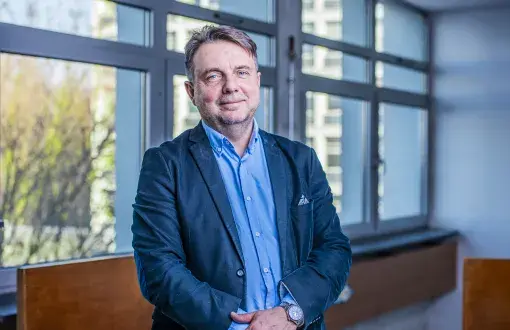 Mężczyzna w granatowej marynarce i błękitnej koszuli stoi na tle ściany z oknami