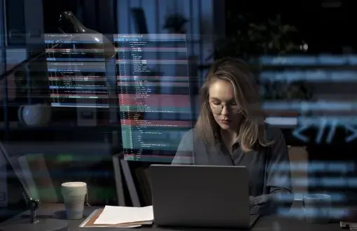 Kobieta pracująca wieczorem przy laptopie w biurze, z okularami na nosie; nad biurkiem widoczne holograficzne wyświetlacze z liniami kodu programistycznego , obok lampka biurkowa, kubki z kawą i dokumenty na blacie.