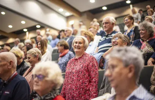 Grupa seniorów siedzi na widowni sali wykładowej. W centrum zdjęcia uśmiechnięta kobieta w czerwonej bluzce w serca, wokół niej pozostali uczestnicy spotkania.