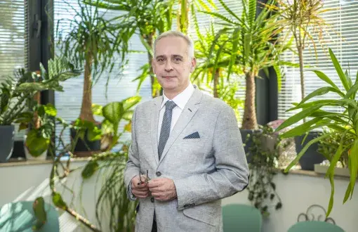 prof. J. Grems w jasnym garniturze stoi na tle pokoju z dużą liczbą zielonych kwiatów doniczkowych