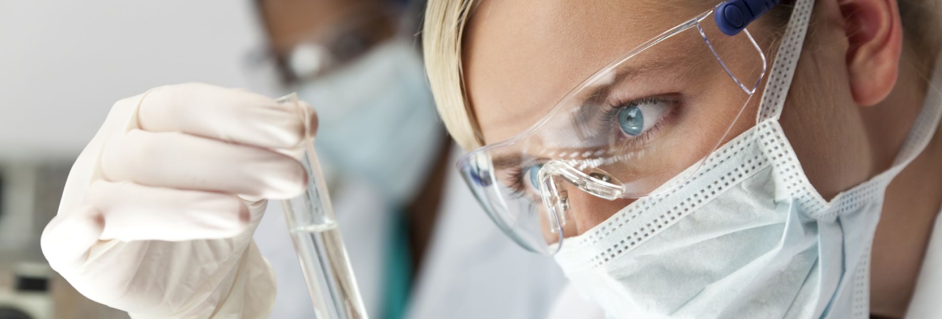 Kobieta naukowiec w białym fartuchu, masce i okularach ochronnych przygląda się probówce.