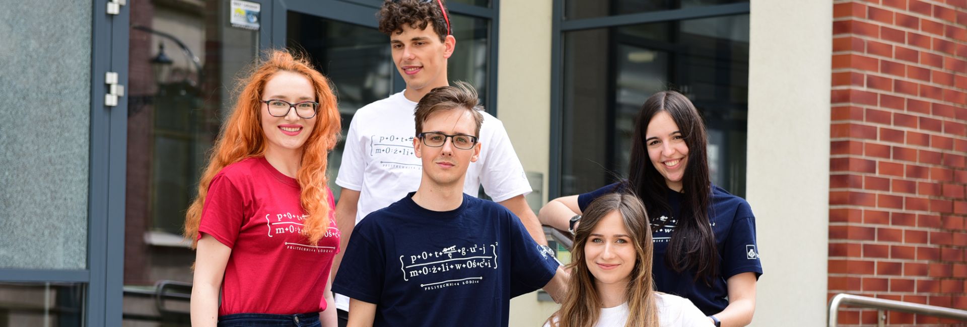 Pięcioro uśmiechniętych studentów w kolorowych koszulkach stoi na schodach przez szklano-ceglastym budynkiem.